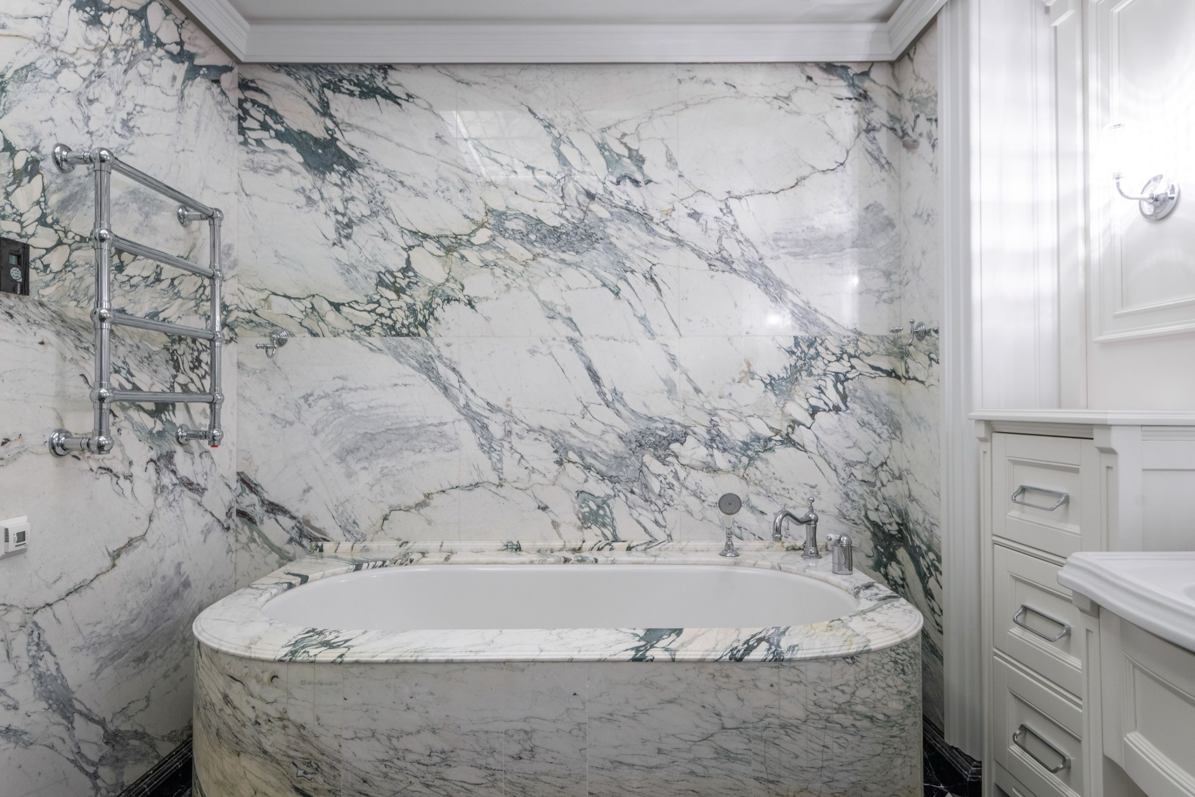 Bañera ovalada revestida del mismo mármol blanco con vetas grises y verdosas que cubre las paredes, grifería y toallero cromados de estilo clásico y un mueble de cajones blanco a la derecha.
