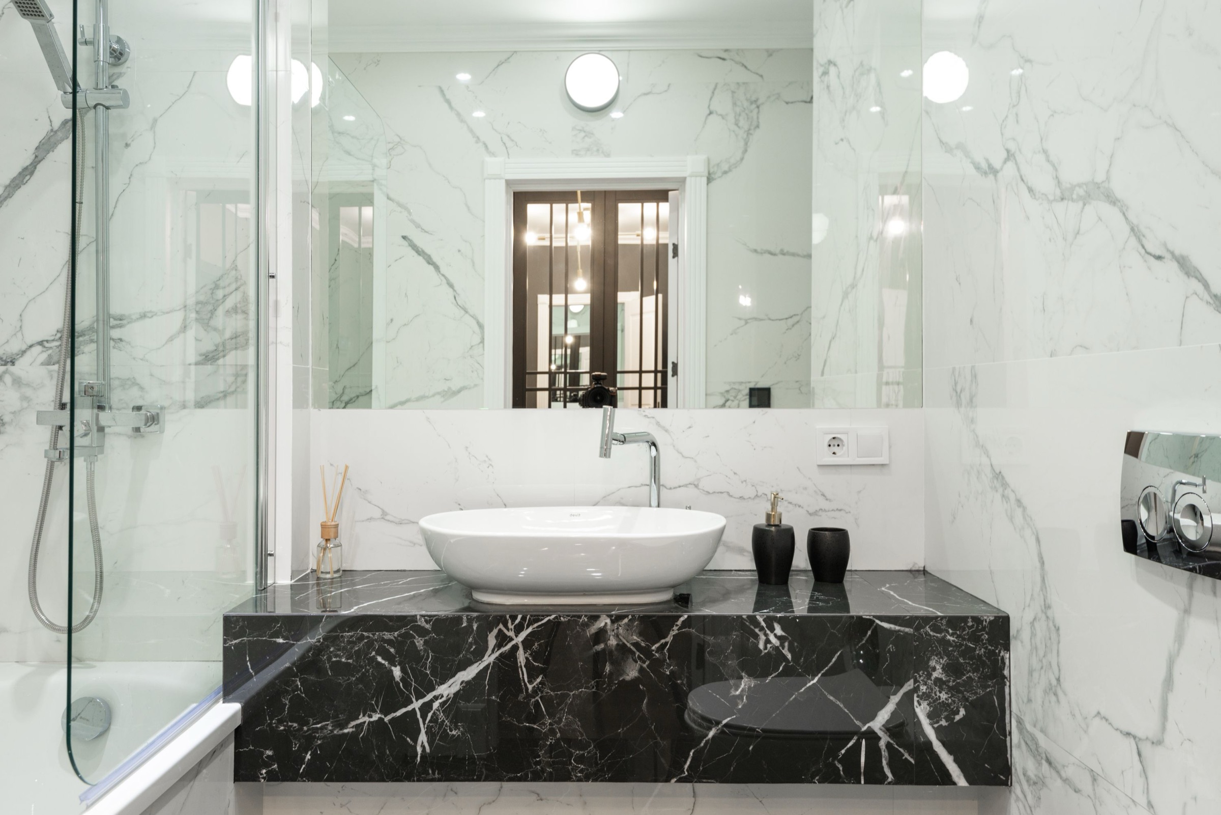 Baño revestido de mármol blanco con vetas grises, lavabo de cerámica blanca sobre una encimera volada de mármol negro con vetas blancas, grifería cromada, gran espejo, mampara de cristal y accesorios negros.