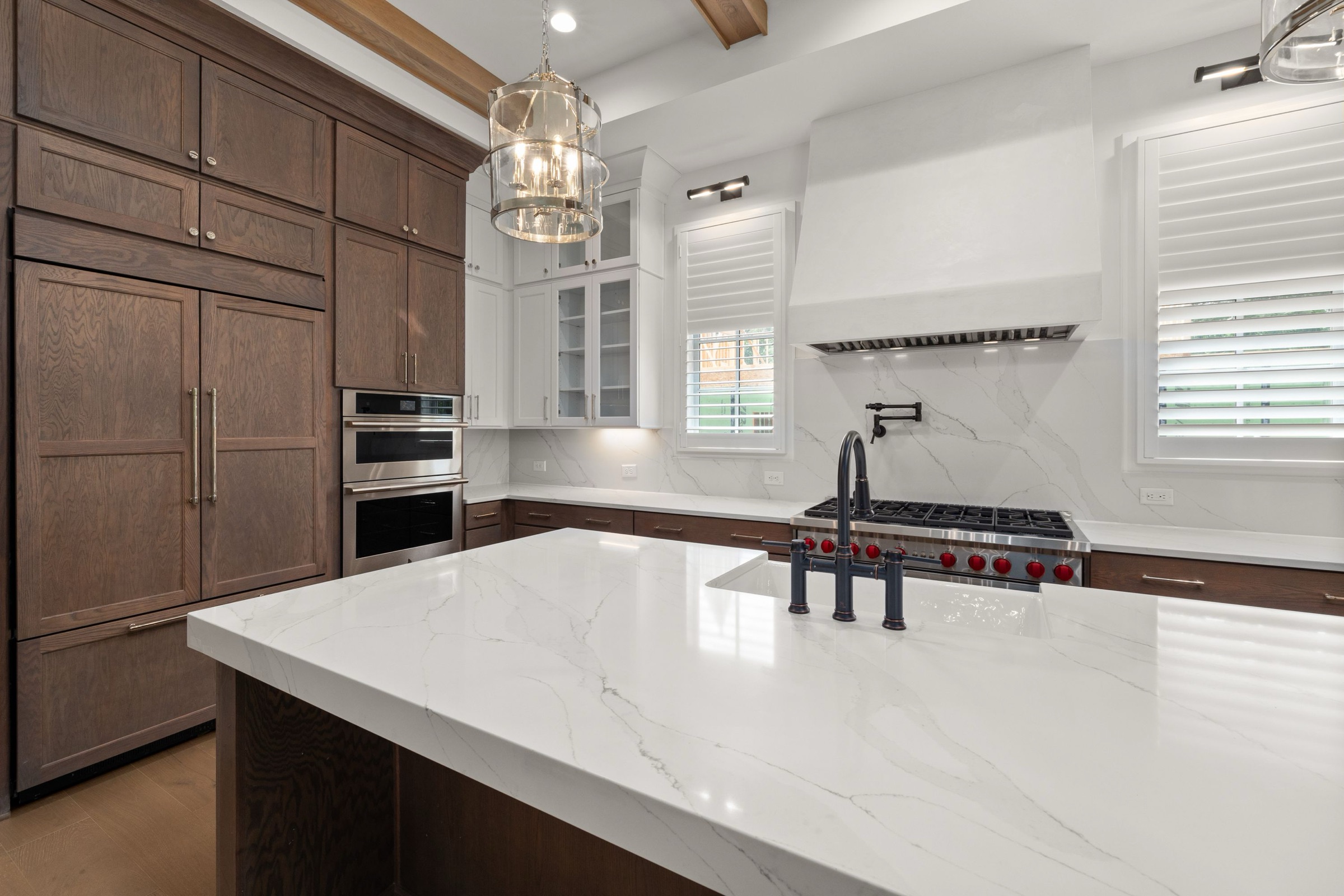 Cocina de diseño con isla de piedra blanca veteada en gris y grifo negro, armarios de madera de roble oscuro, frente de la misma piedra, cocina profesional de acero con mandos rojos, campana blanca de obra y lámparas colgantes de cristal.