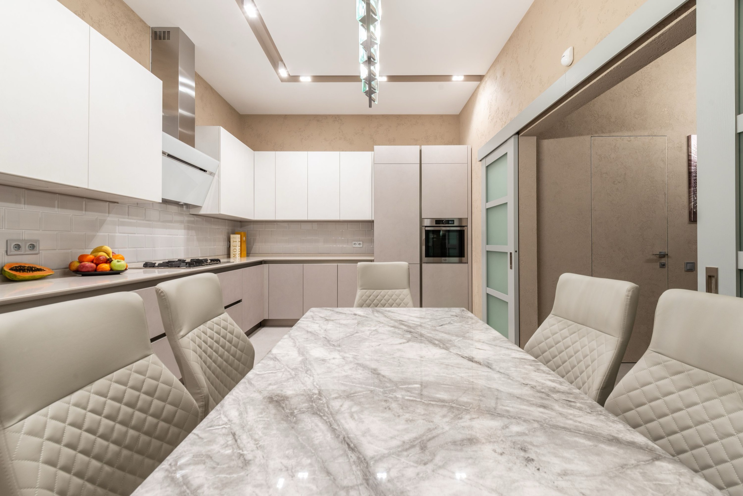 Cocina contemporánea en blanco y beige vista desde la cabecera de una mesa larga de mármol gris pulido, rodeada de sillas tapizadas acolchadas; al fondo, muebles lacados sin tiradores, horno integrado y una puerta corredera de cristal.