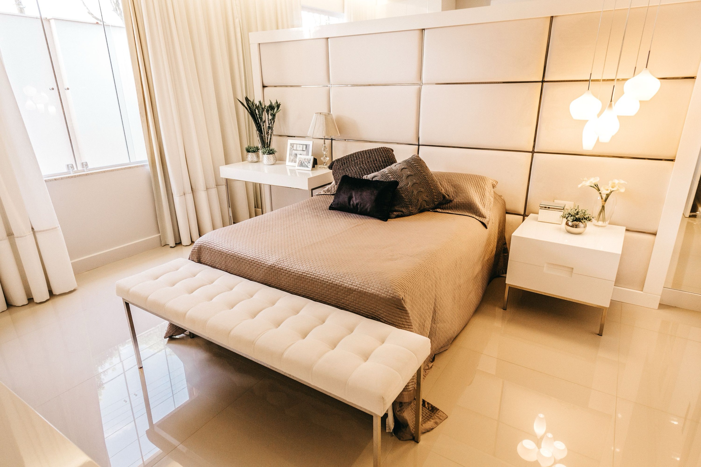 Dormitorio en tonos crema con pared de cabecero tapizada en paneles, colcha color topo con cojines, banco capitoné blanco a los pies de la cama, mesillas lacadas blancas, lámparas colgantes de cristal, cortinas de lino y suelo brillante.