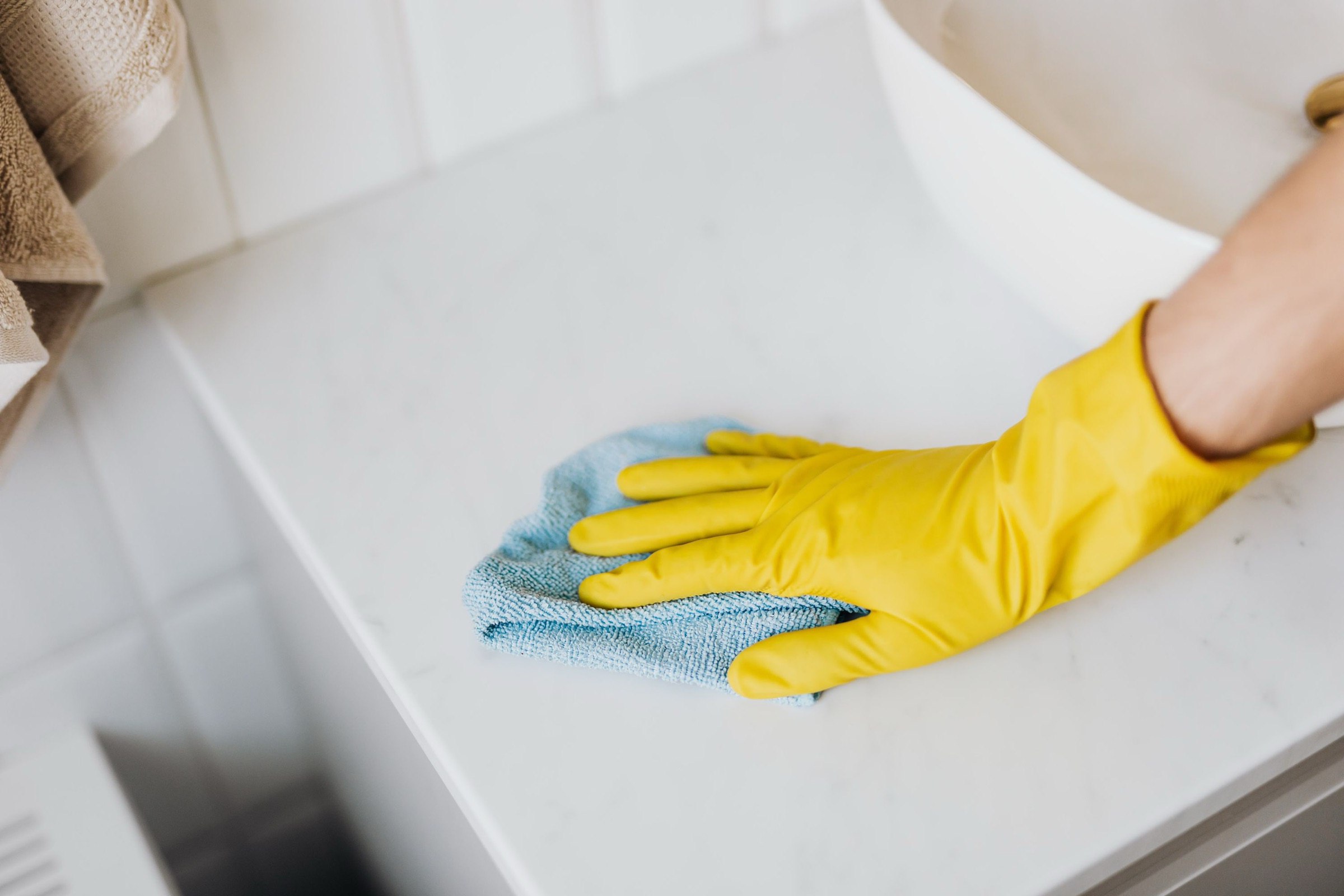 Mano con guante de goma amarillo limpiando con un paño de microfibra azul claro la encimera blanca de un baño, junto al borde de un lavabo y una toalla beige, con luz natural suave.