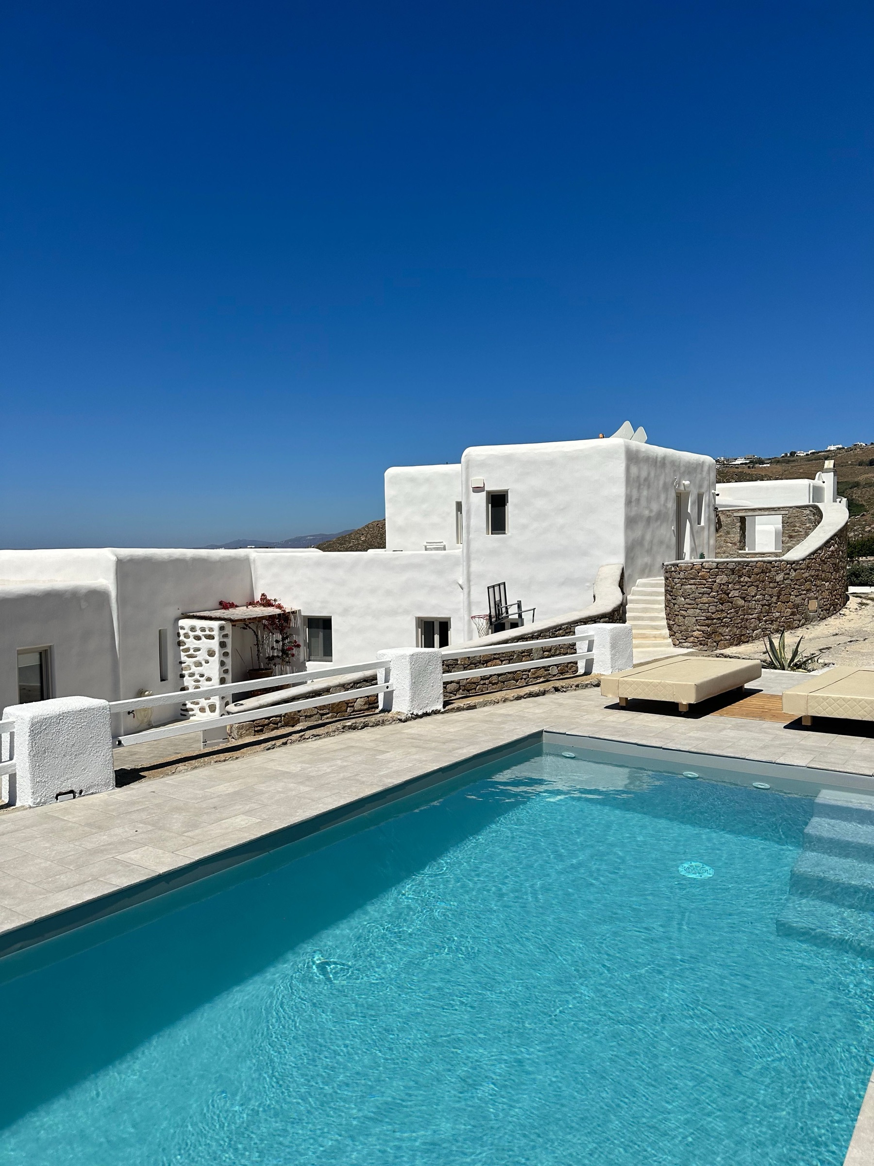 Piscina de agua turquesa con suelo de piedra clara frente a una villa encalada de volúmenes blancos, muros de piedra seca y tumbonas, bajo un cielo azul intenso sin nubes.