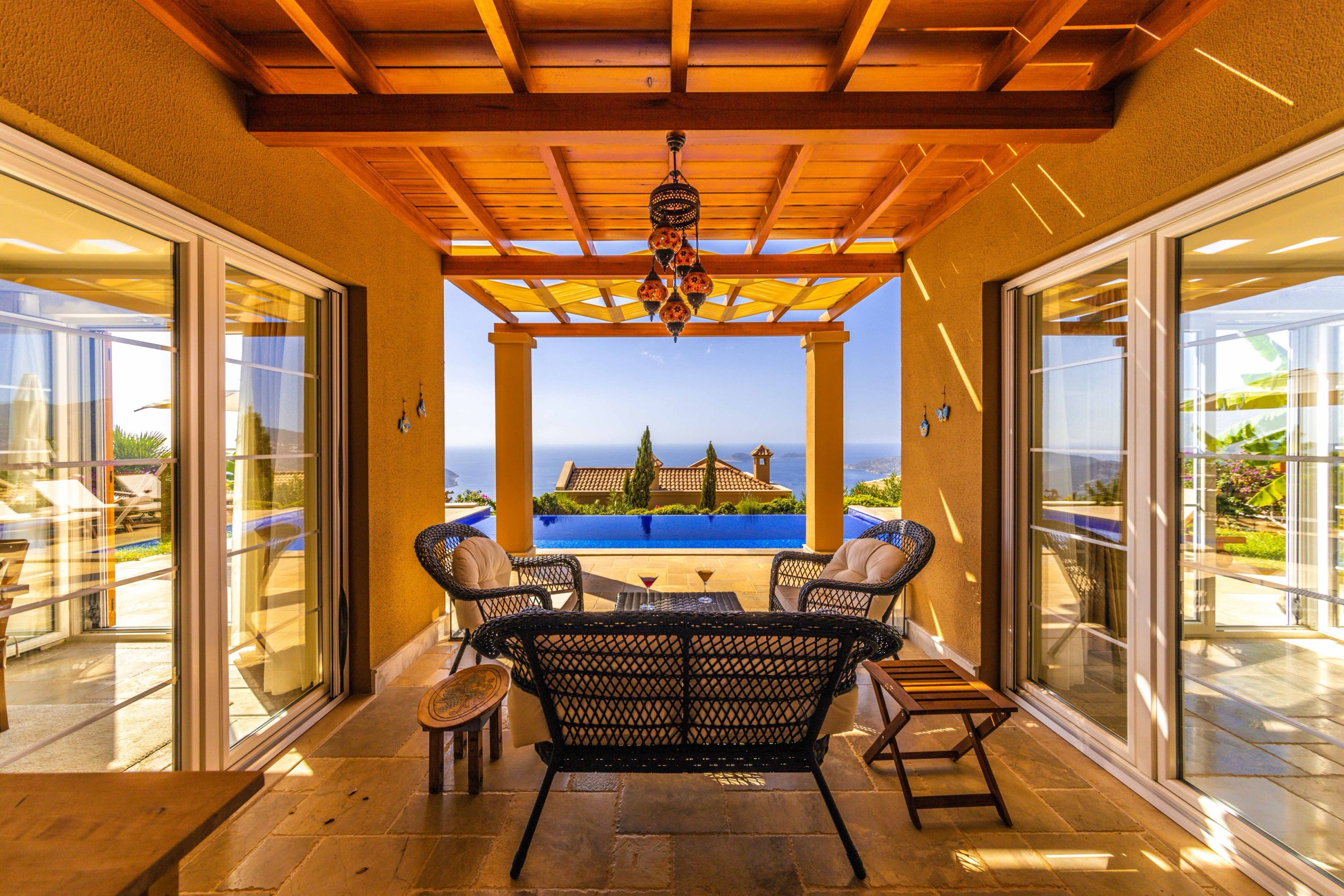 Porche de una villa con techo de vigas de madera, suelo de piedra natural y muebles de ratán negro, abierto entre dos puertas acristaladas hacia una piscina desbordante, cipreses y el mar azul al fondo, con luz cálida de tarde.